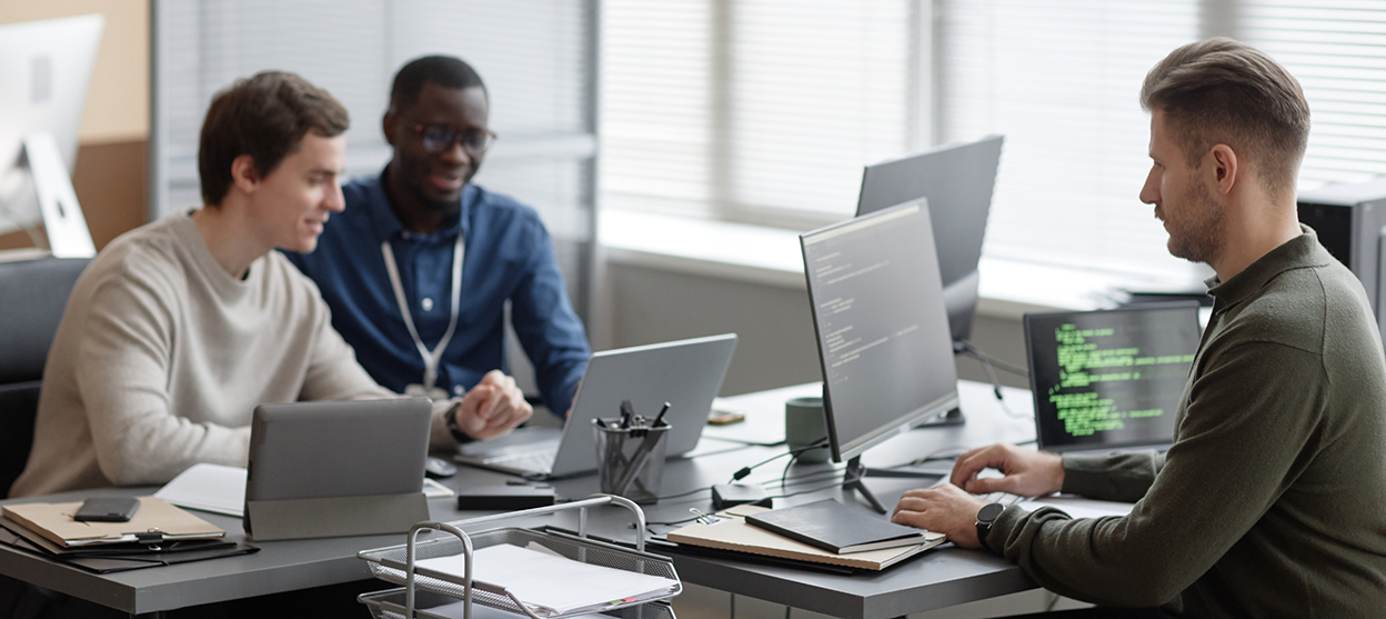 1 main - VIRTUTEM Three developers collaborate at a desk with laptops and code visible on monitors in a bright office.