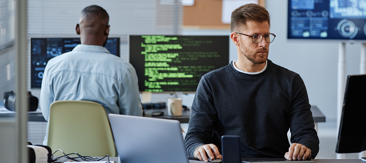 1 main - VIRTUTEM Software developers in a modern office; one man with glasses types on a laptop while code shows on a monitor nearby.