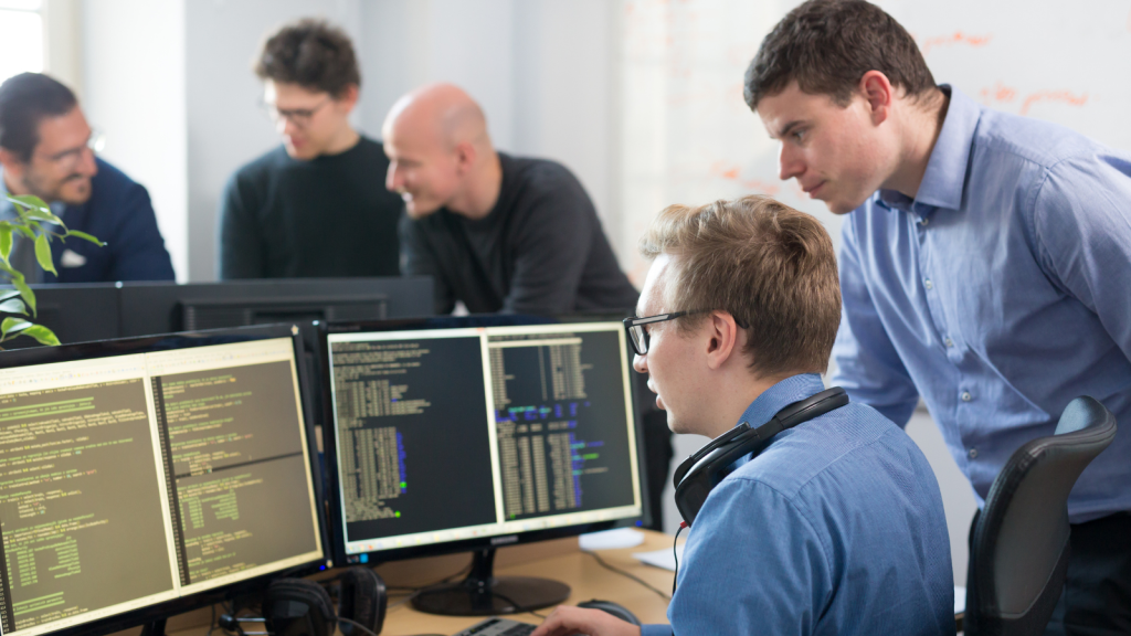 Four developers collaborate around a desk with multiple monitors showing code; one seated with headphones reviewing code while teammates discuss nearby.