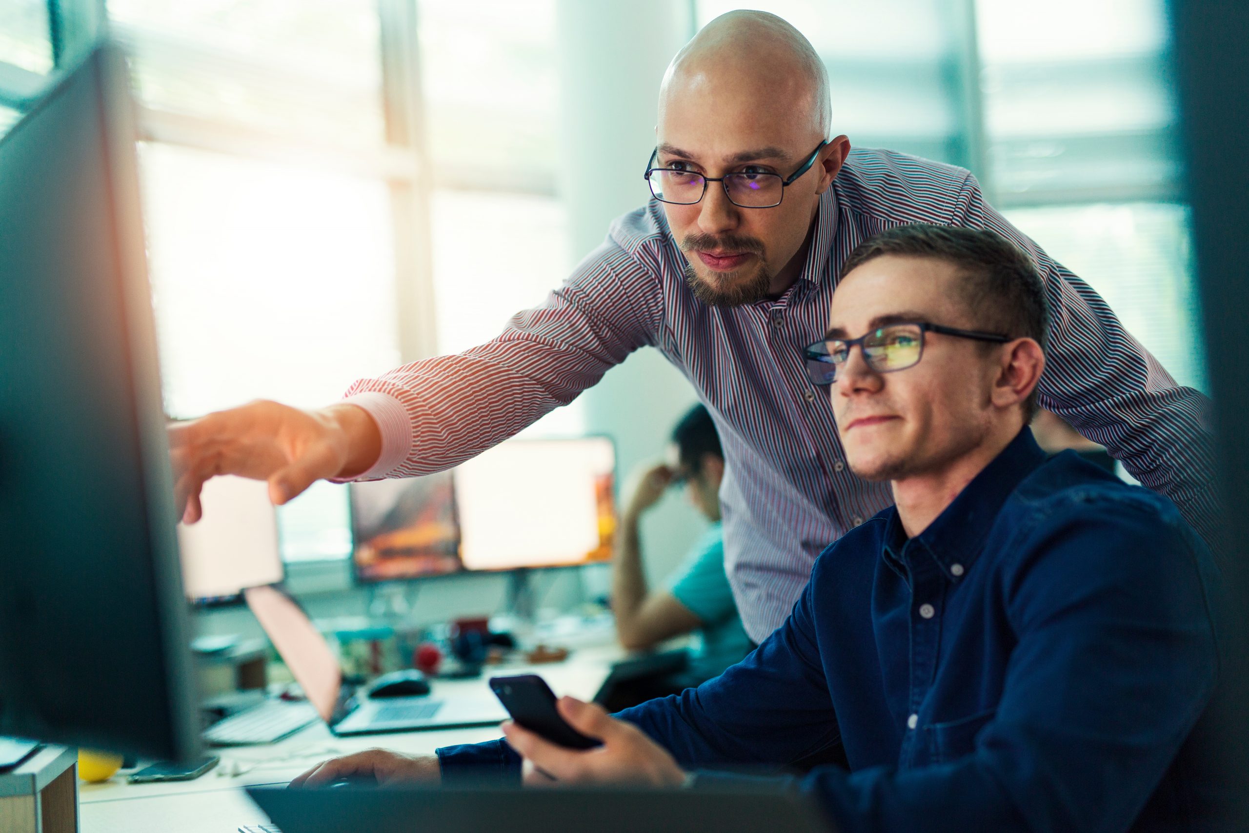 Senior developer pointing at a monitor while guiding a colleague at his desk